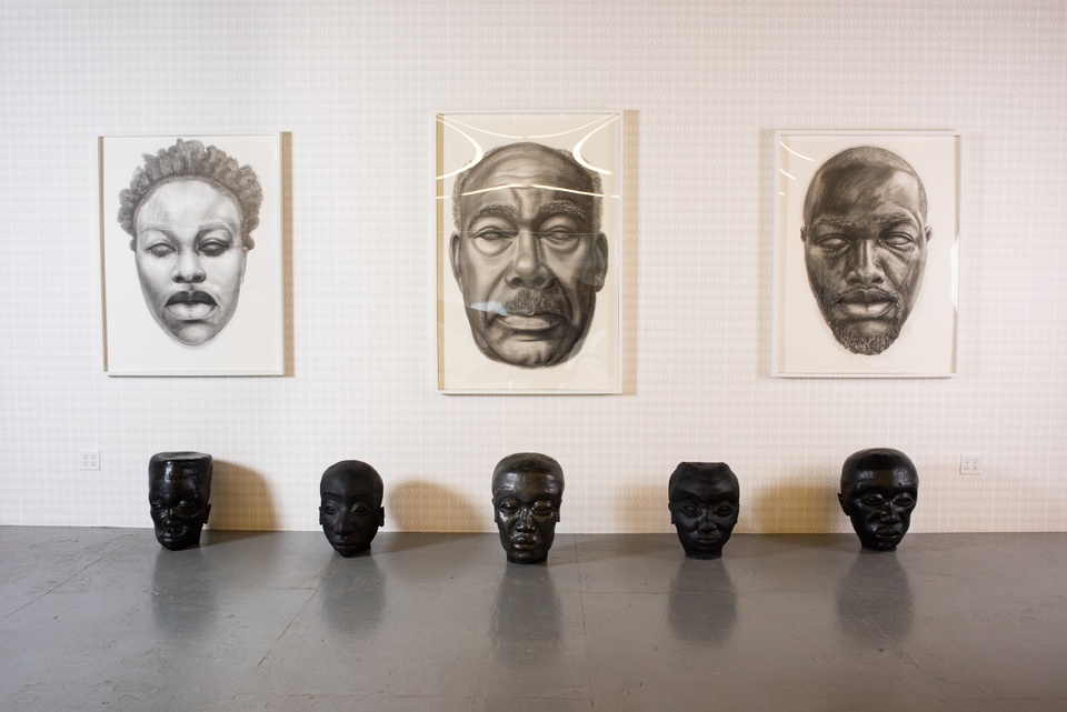 Three large-scale black and white drawings of heads with blank pupil-less eyes and a row of five black sculptural heads sitting on the floor below.