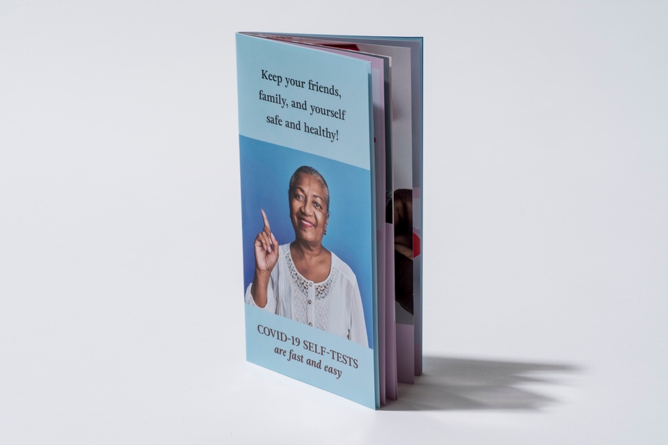 Standing blue booklet with a picture of an old woman on the front reads, "Keep your friends, family, and yourself safe and healthy! COVID-19 Self-Tests are fast and easy."