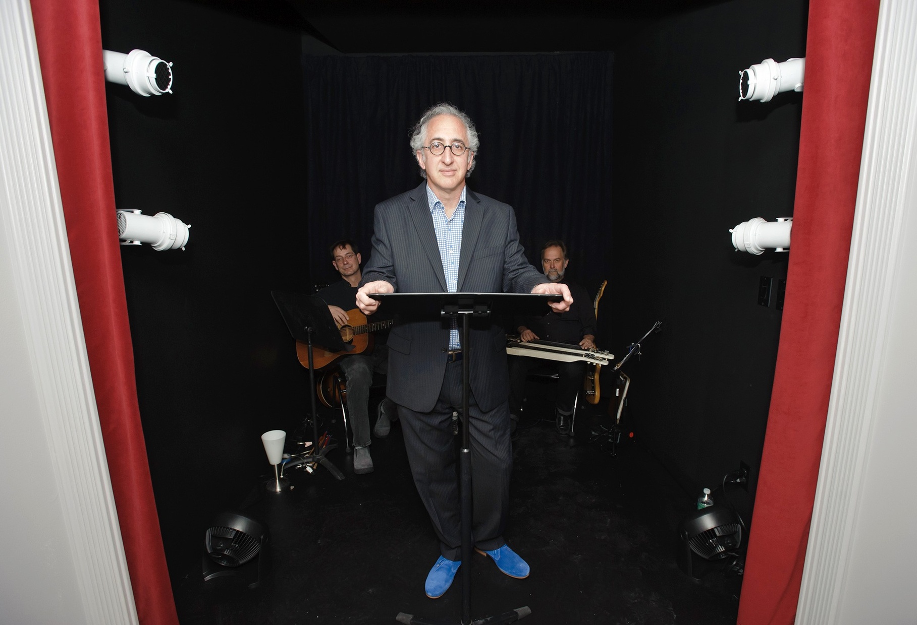 An older, tanned man stands behind a music stand in a black room with two men sitting behind him holding stringed instruments.