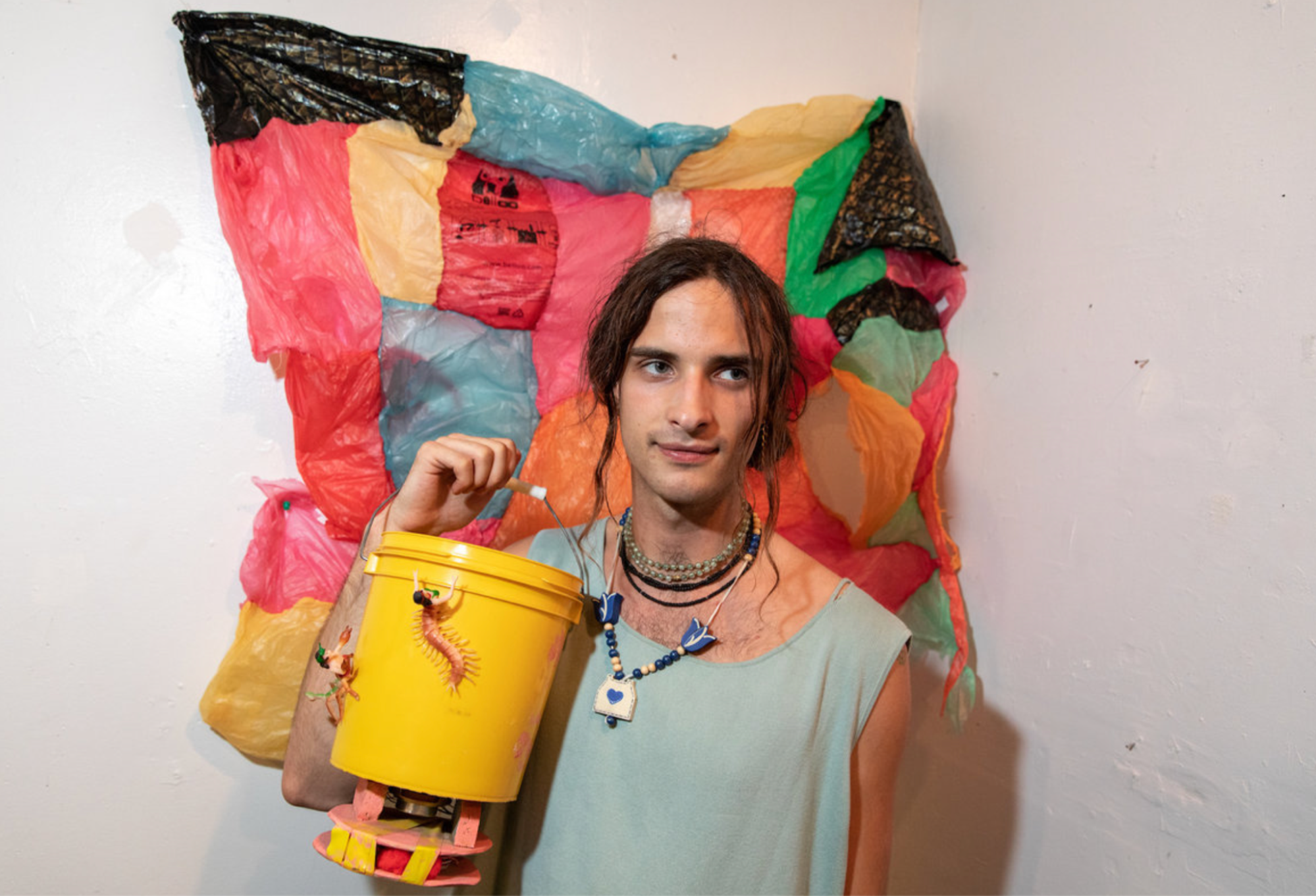 A person with long hair pulled back with strands hanging on either side of their face holds up a yellow bucket at chest level. Behind them is an artwork that looks like a multicolored patchwork quilt made of plastic bags.
