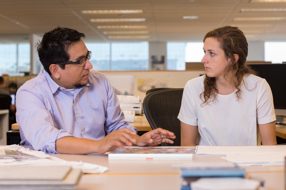 Two people sit on the same side of a table and talk.