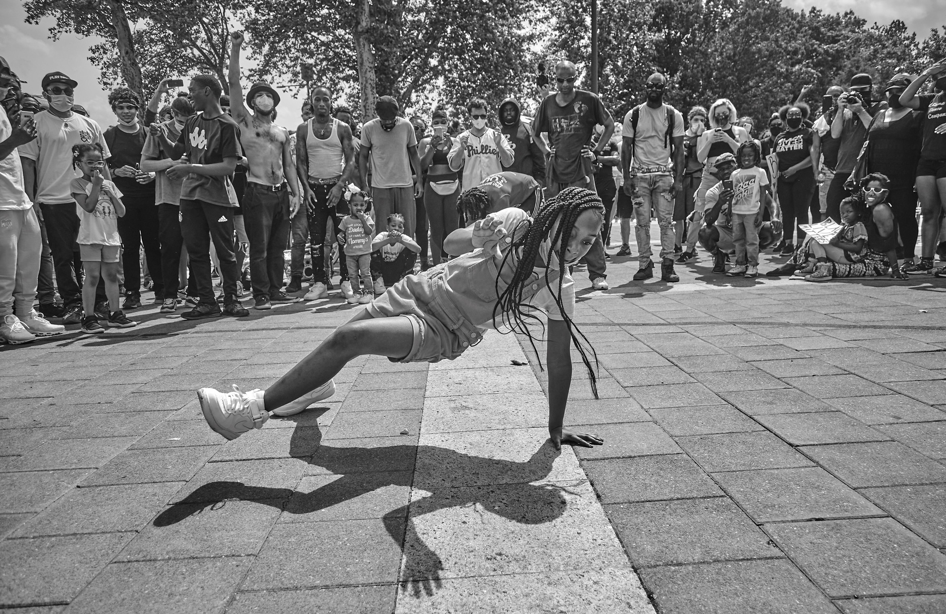 A black and white photograph of a young, Black girl dancing surrounded by a circle of people looking on and cheering.
