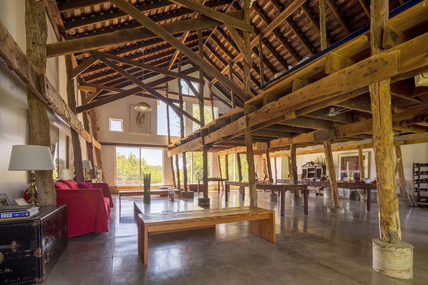 Interior of a barn with rough, exposed wooden beams with a loft and a gabled roof. The floor is shiny concrete and large windows have been added to the back wall.