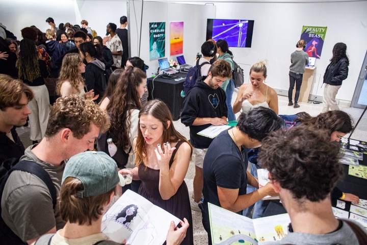 Crowded gallery space with posters on the wall representing illustration and design projects. People flip through picture books and watch animations on screens around the room.