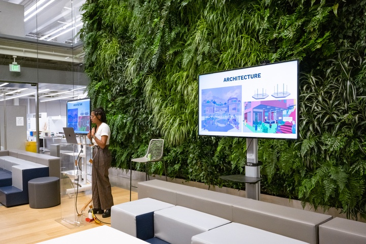 Hillawi Abraham stands at a lecturn in front of a living green wall. Two video screens behind her read "Architecture" and display heavily saturated renderings of public spaces.