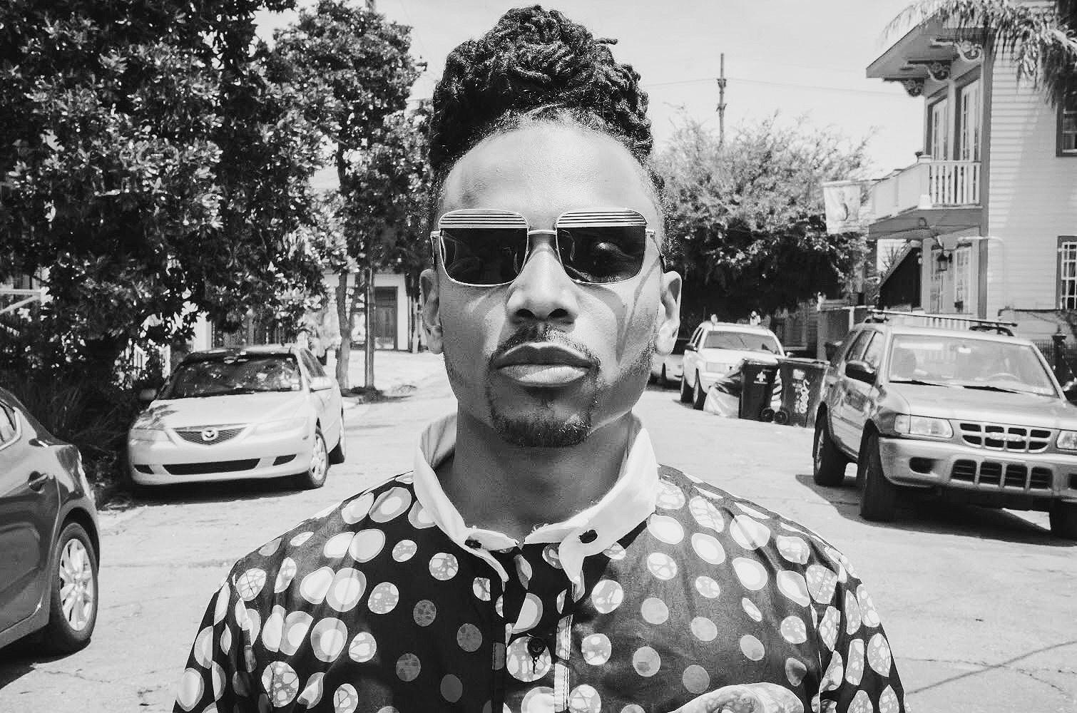 A black-and-white photo of Asante Amin standing in the middle of a street lined with parked cars and a tree and two-story building in the background. Amin wears square-framed metal sunglasses, a shirt patterned with a design of small circles, and squarely faces the camera. 