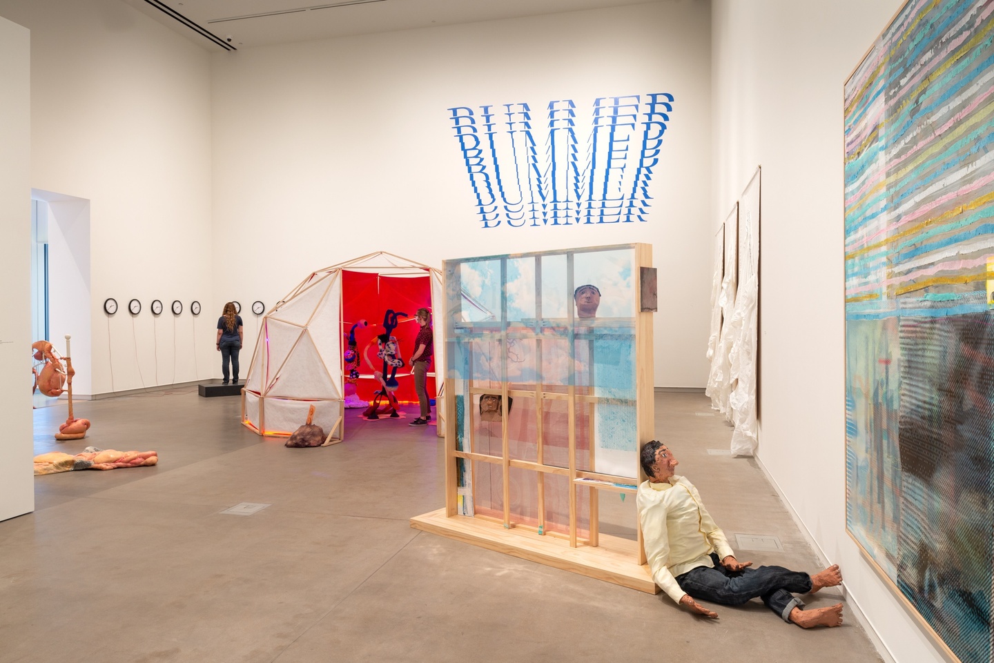 Installation view of artworks at Kemper Art Museum, including several clocks on the back side wall, a dome-like structure of white canvas and wooden pieces, opened to a bright reddish-pink interior with a person standing inside; the words "Bummer" written over and over in blue high on the back wall; a wooden, window-like sculpture with a 3D figure of a person sitting, leaned against it; and two pieces on the side wall, one in all white, the other featuring multi-colored horizontal stripes.