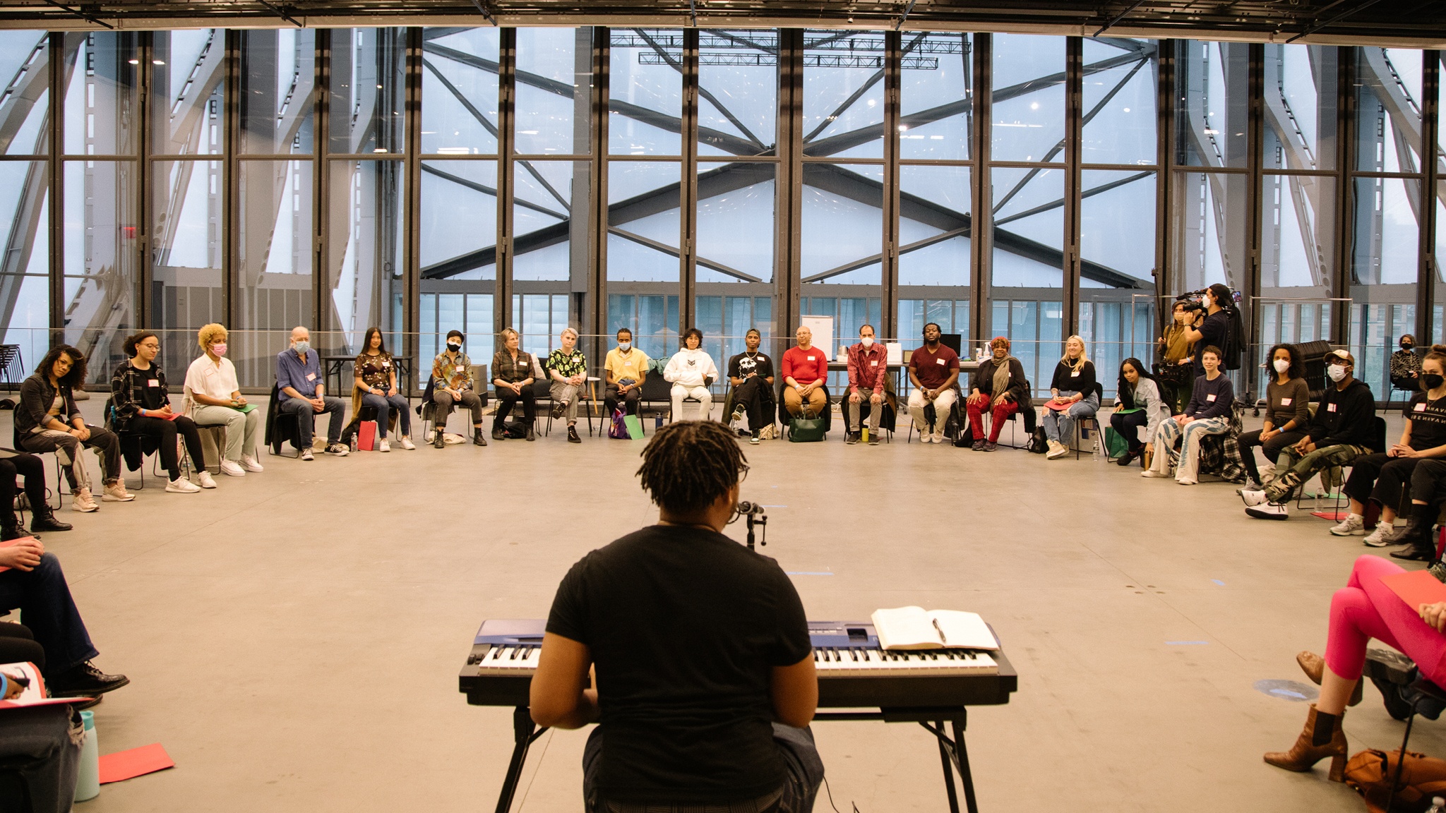 The artist Troy Anthony sits at a keyboard with his back to the camera facing a semicircle of people of various ages and races seated across a wide performance space with high ceilings at The Shed
