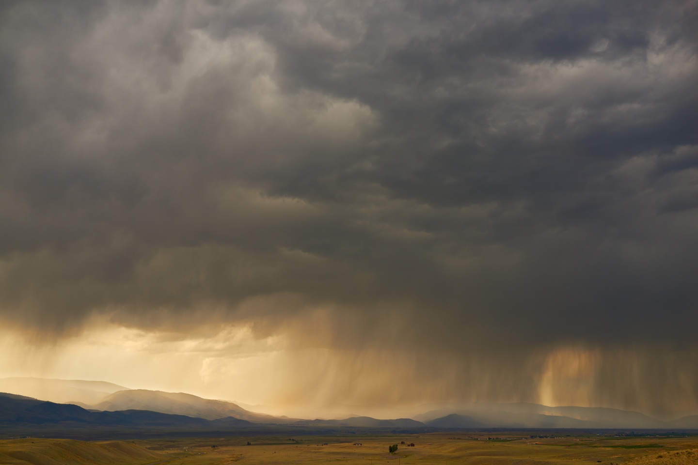 Landscape photo of a dense bank of rainclouds taking up 2/3 of the image, over a sandy plain climbing to foothills in the distance.
