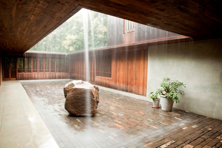 Courtyard area, with light streaming through a rectangular opening in the roof. The courtyard featured a larger, rectangular inset of brick pavers, with a larger stone boulder and a couple of green ferns. Wood walls line the back and part of the right side of the walls, and trees are visible through the roof opening.