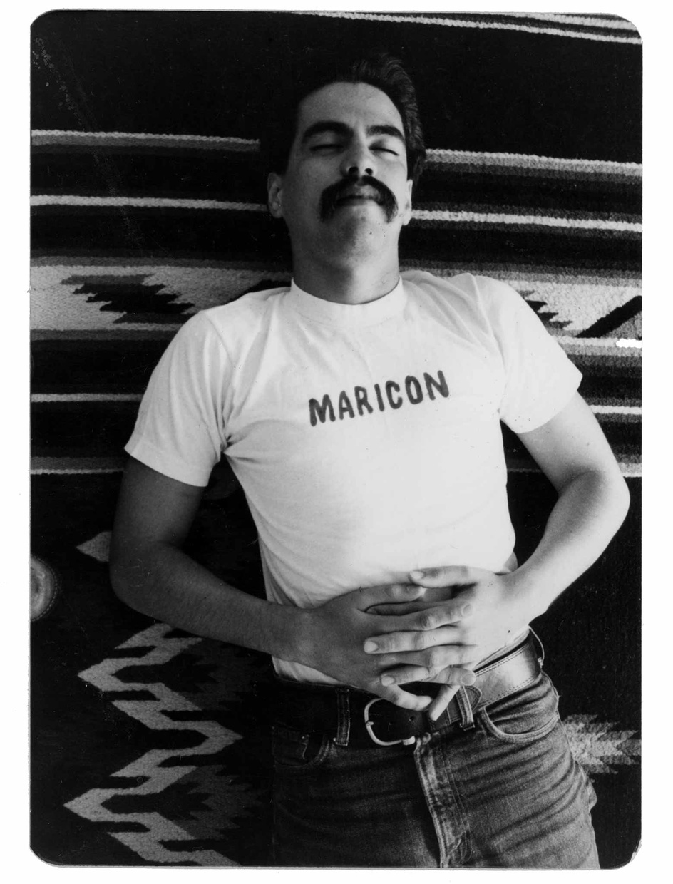 A black and white photo of a Latino man laying on a blanket wearing a shirt that says "MARICON."