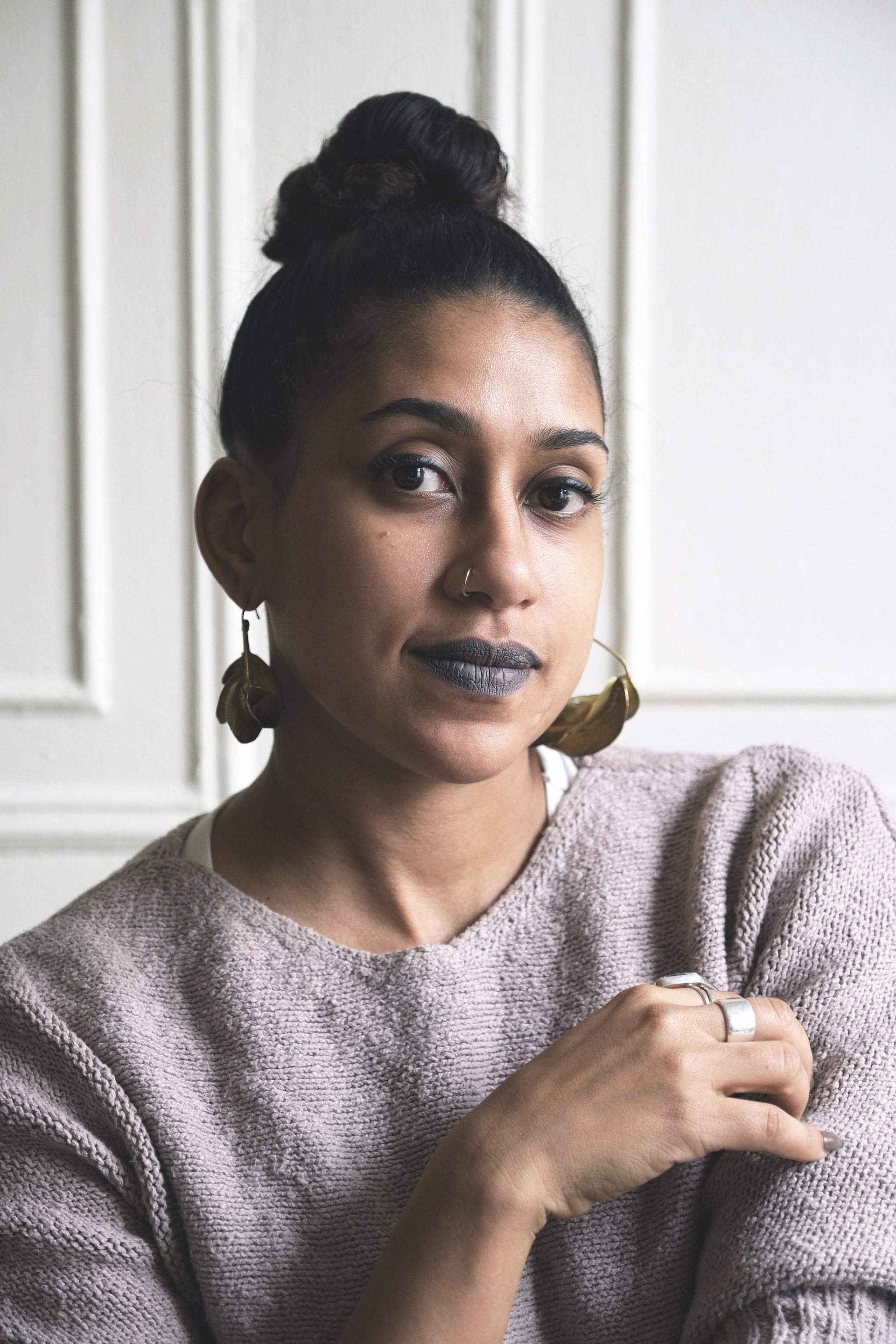 A photo of artist Shani Peters. She wears a pale lavender sweater and sits with one hand drawn up to her left shoulder. She wears gray lipstick and has her hair pulled into a loose bun on top of head. 