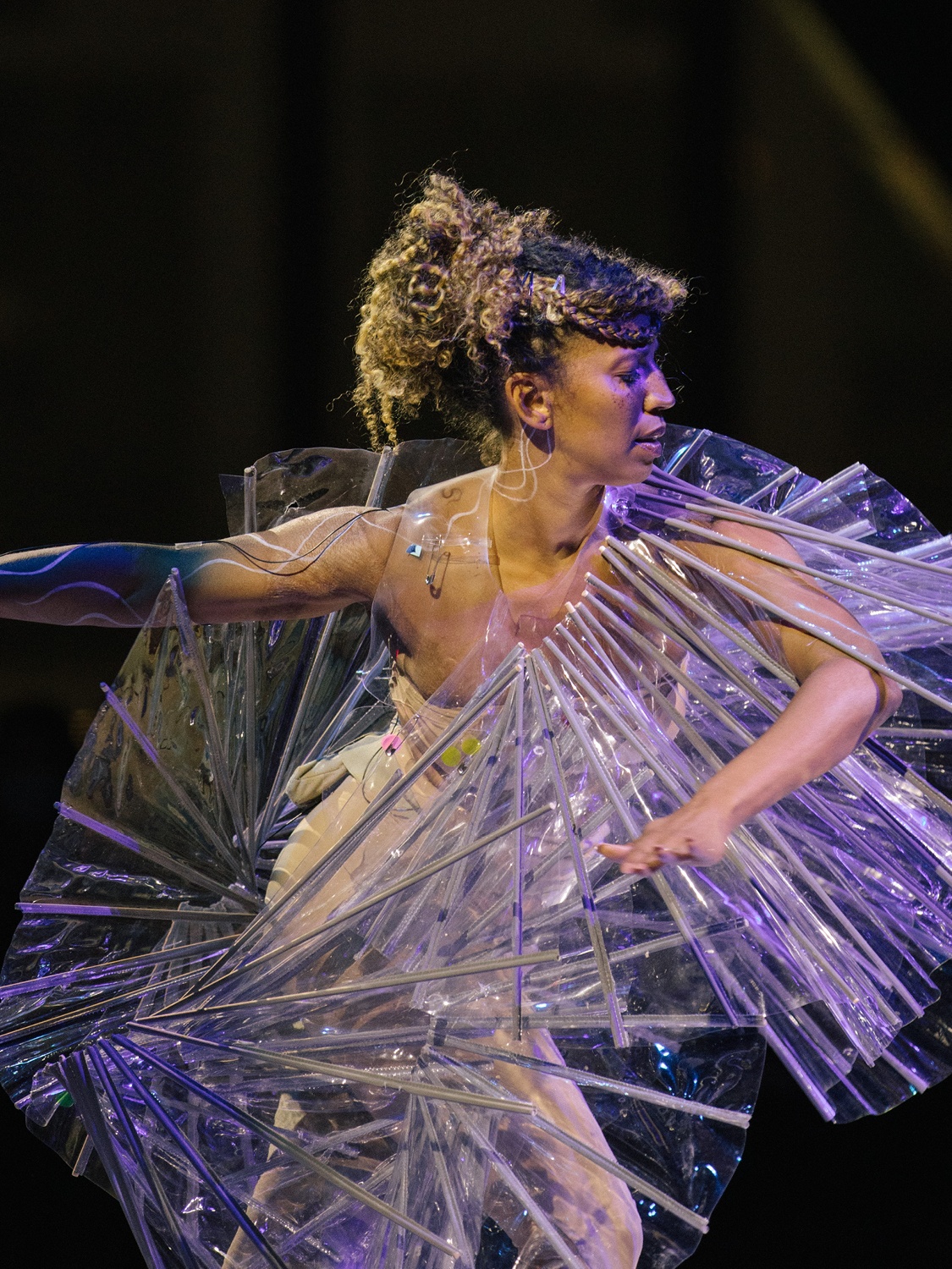 A performer in a sculptural costume holds arms out while lit by a stage spotlight