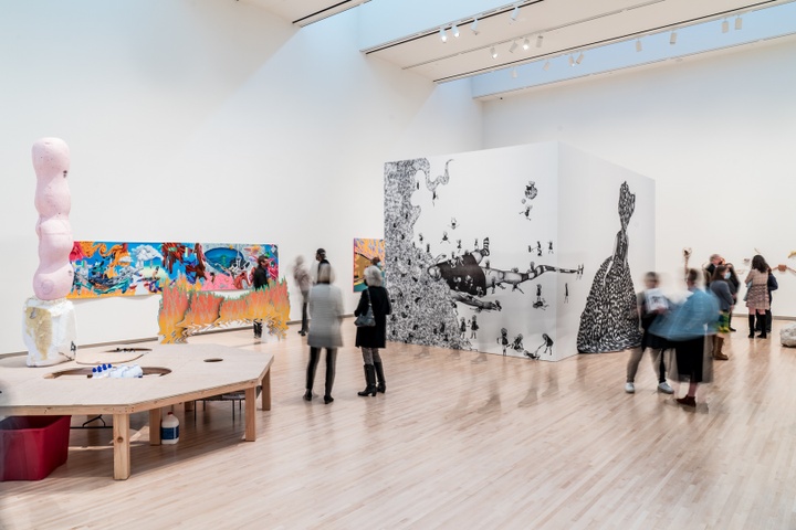 Wide view of a museum gallery space displaying a wooden stage installation, a brightly-colored painting on the back wall, and a half-wall in the center of the gallery wrapped in a large-scale print of a black and white drawing of women with pouffy afro hair.