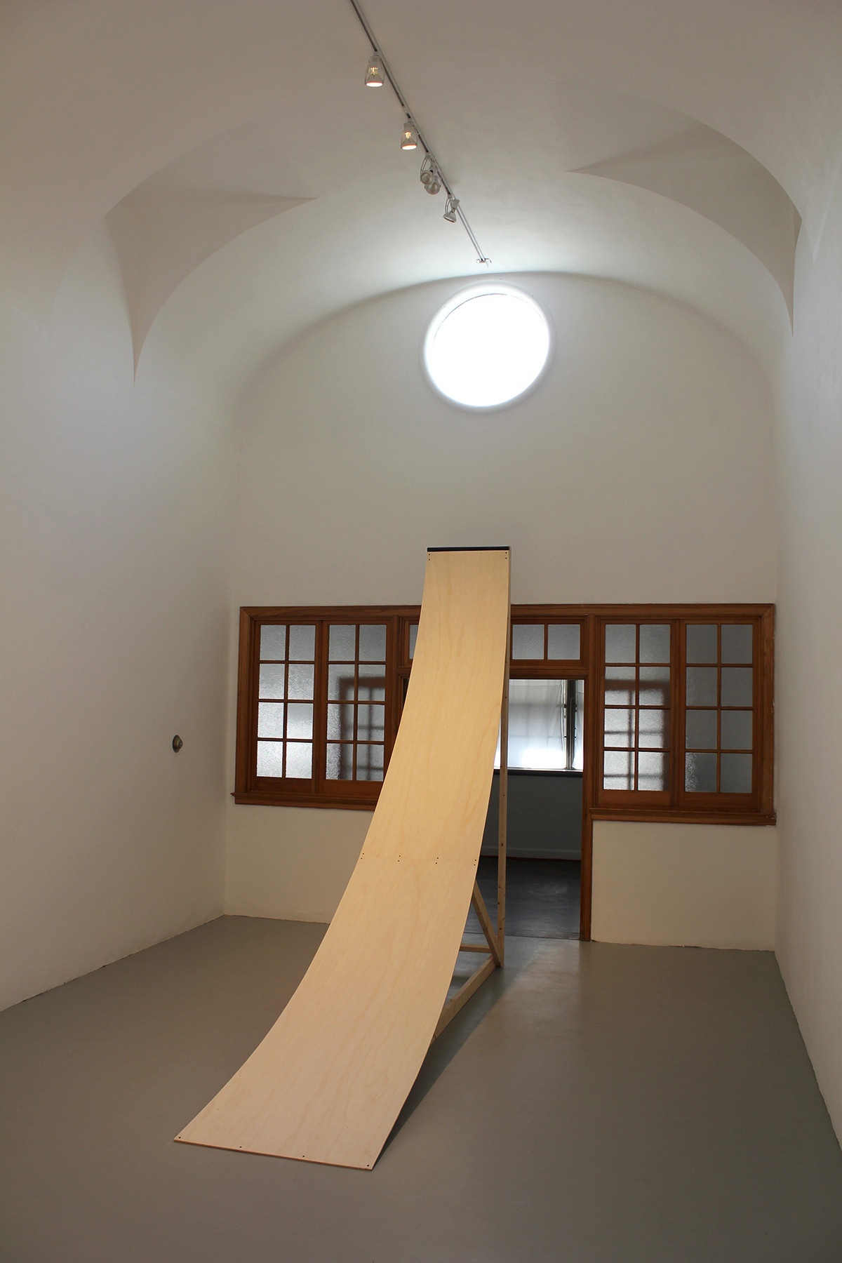 A gallery space: the walls are white and arch overhead, the floor gray. There is a circular window at the top in the center, through which bright light shines through, illuminating the gallery: in the center of the space is a curved wooden ramp supported by a wooden structure in front of an opening (whose windows and door are framed with wood).