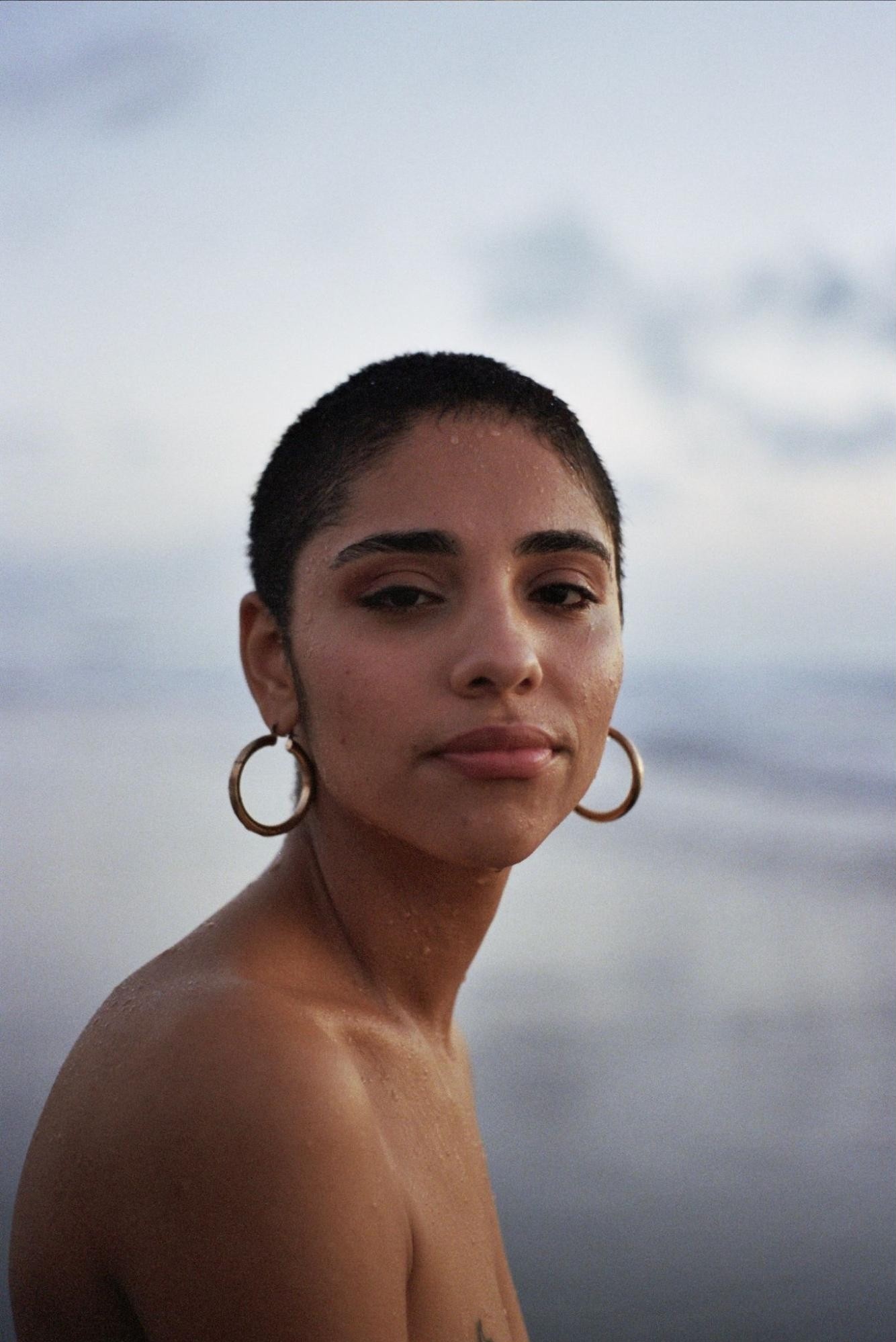 A Dominican American woman with short hair seen from the shoulders up. Her shoulders are bare, she wears gold hoop earrings, and is wet as if she has just been bathing in a body of water. In the background is a hazy sky.