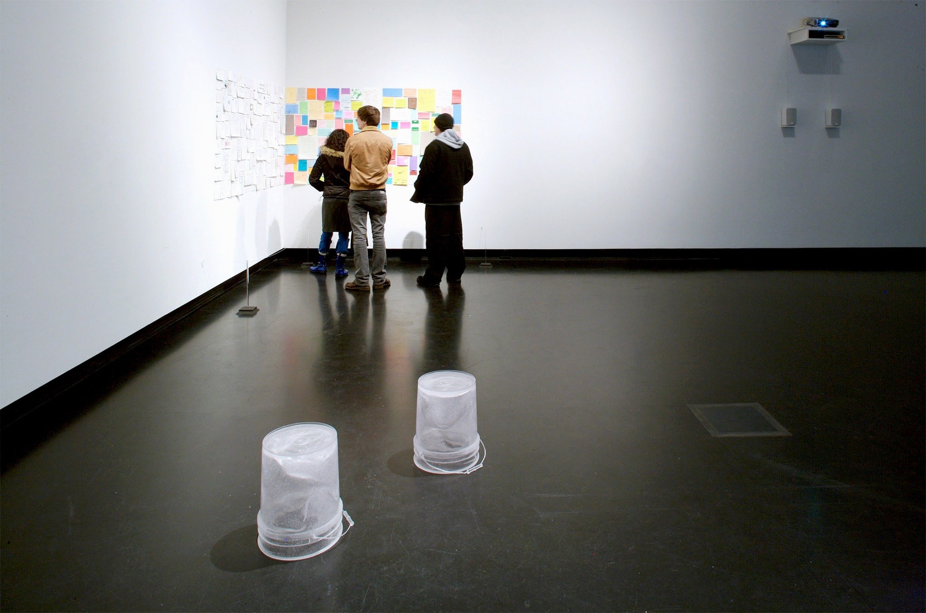 A group of three people stand looking at an artwork hanging on a white wall made of different colored, different sized squares and rectangles. On the floor in front of them, two clear garbage pails rest upside-down.