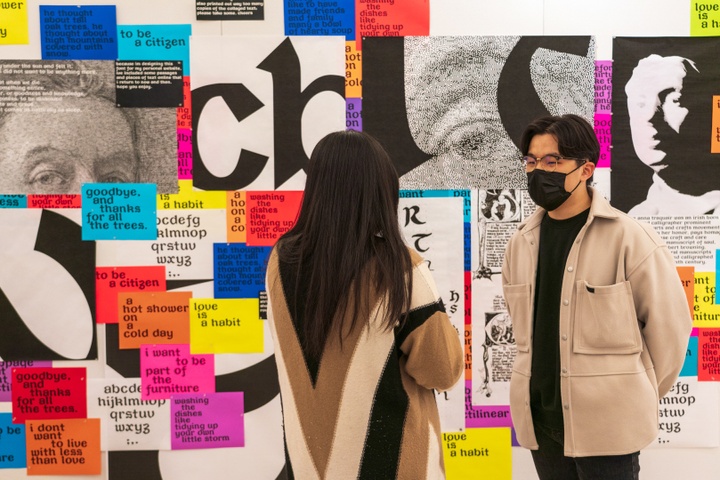 Two people stand in front of a wall covered in brightly colored prints displaying a blackletter font at both small and large scales.