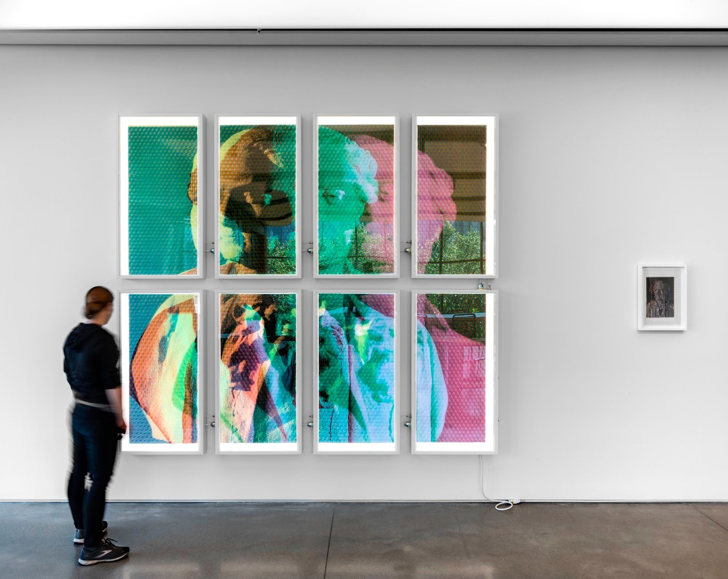 A person standing in front of an artwork made from three photographs converted into red, green, and blue monochrome tonal scales respectively, ultrachrome printed onto translucent, archival plasticized paper, cut by hand into 15mm-wide strips and triaxial woven together by hand. Backlit with eight custom-built 4000K dimmable fixtures.