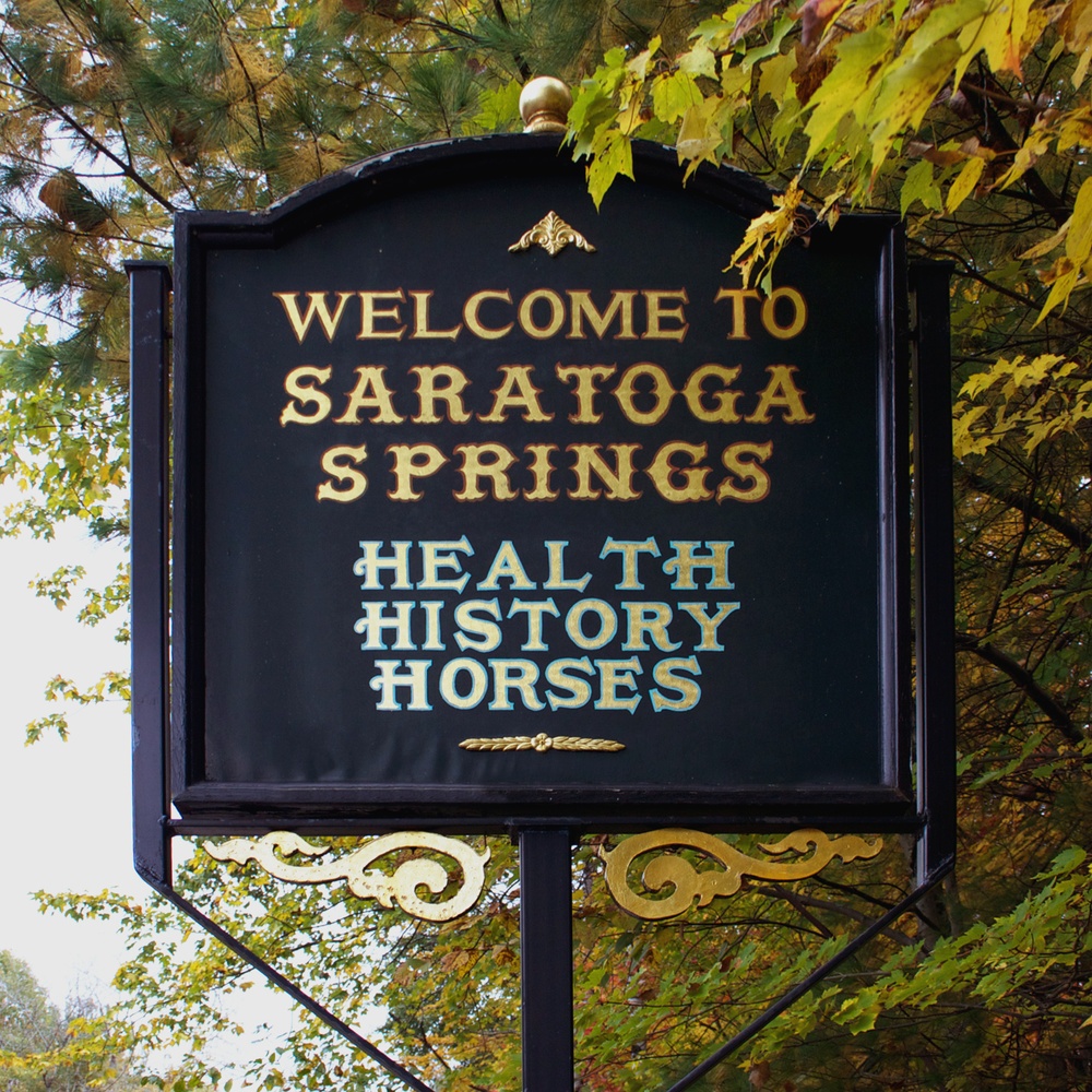 A black sign surrounded by leaves with text "WELCOME TO SARATOGA SPRINGS HEALTH HISTORY HORSES."