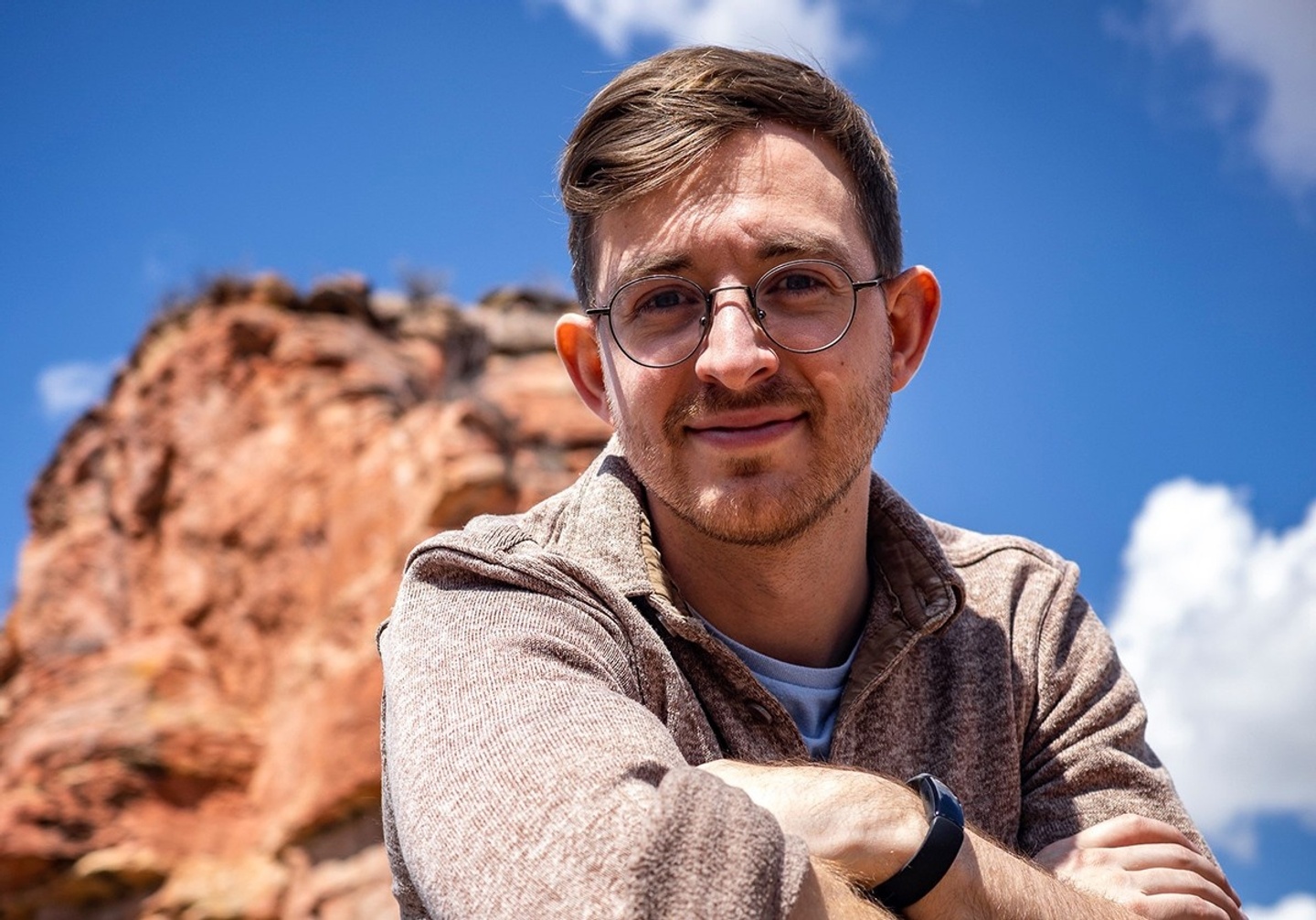 photo of man kneeling down with arms crossed and an elbow resting on one knee. Behind him is mountain and blue skies