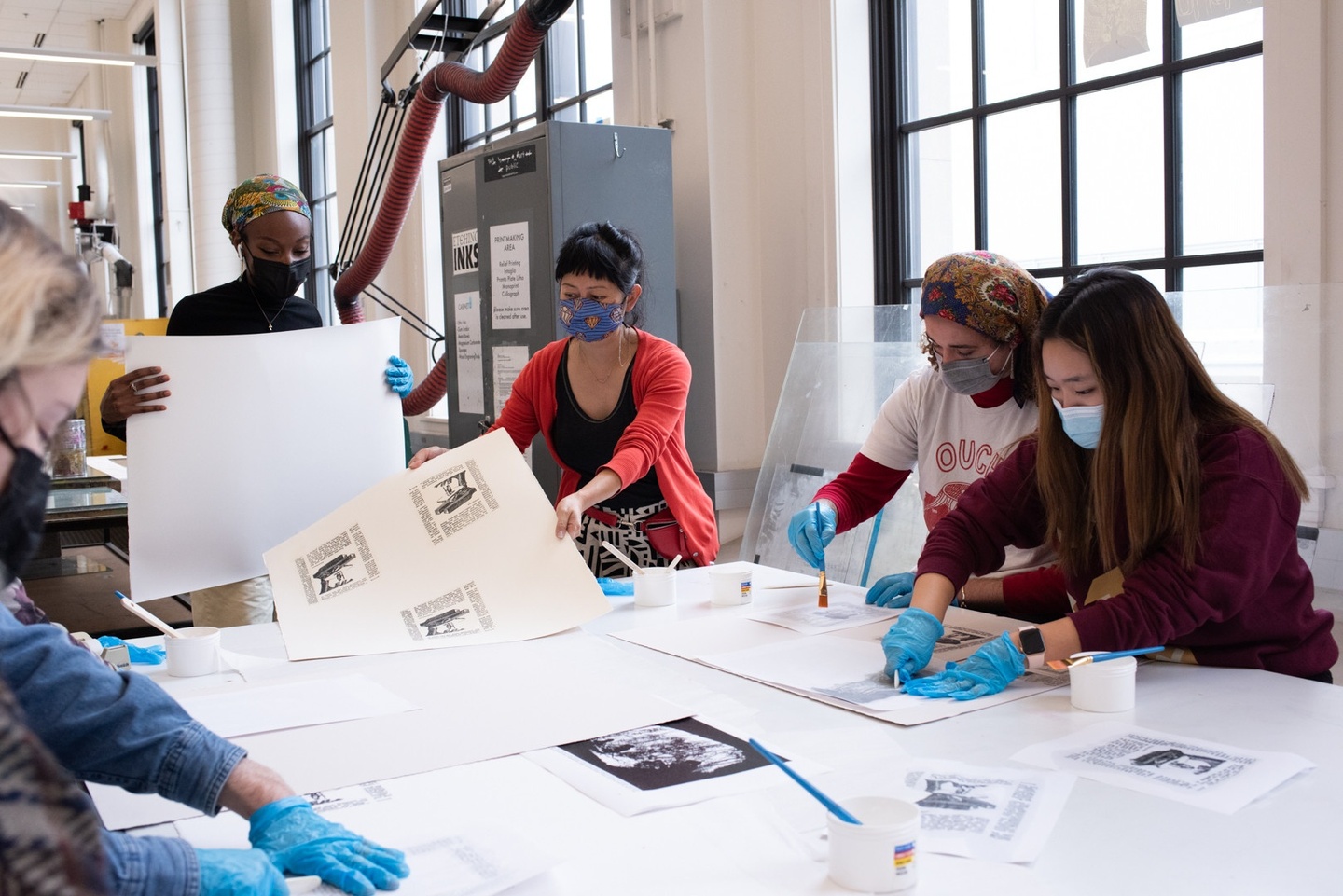 Five or six people gather around a worktable in a printmaking shop, shuffling sheets with artwork around.