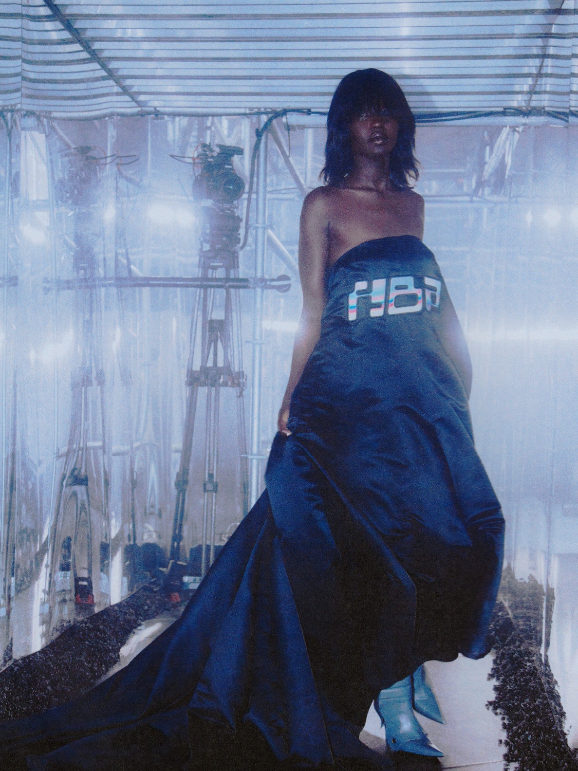 A model in a long black, shoulderless dress stands in front of a scaffolding structure draped in plastic and lit with bright white lights.