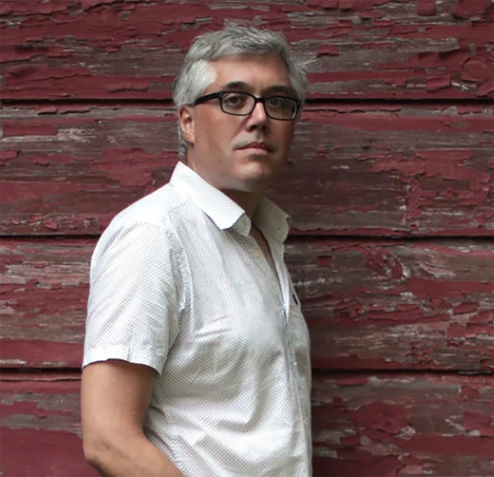 A photo of Brian Rogers, who wears a white, short-sleeve button-down shirt and black-frame glasses. Rogers stands with one shoulder against a wall of wooden planks with peeling red paint. 