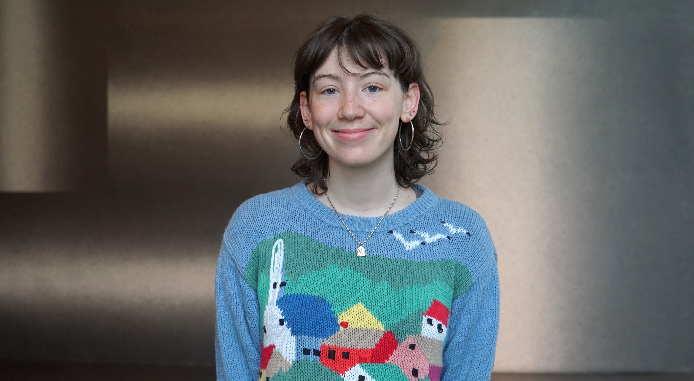 A young light-skinned woman with brown hair smiles at us and wears a light blue, red, and green sweater.