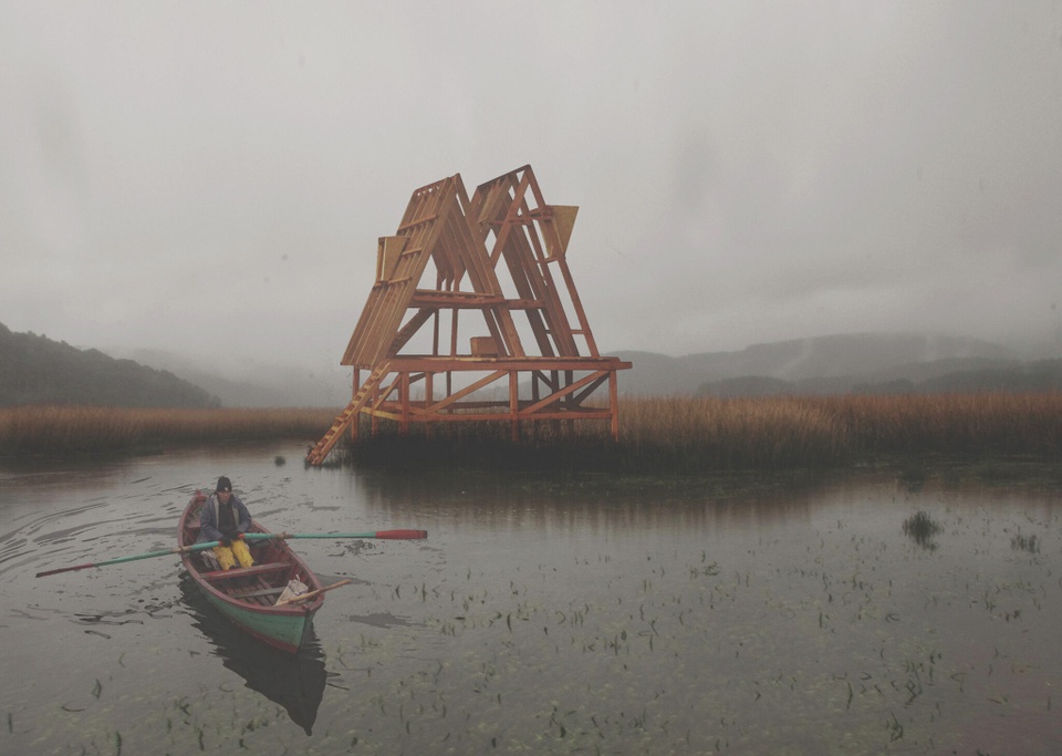 A rendering of a man, paddling backwards in a rowboat in shappow water toward a triangular wooden structure, with tawny reeds and mountains in the misty distance.