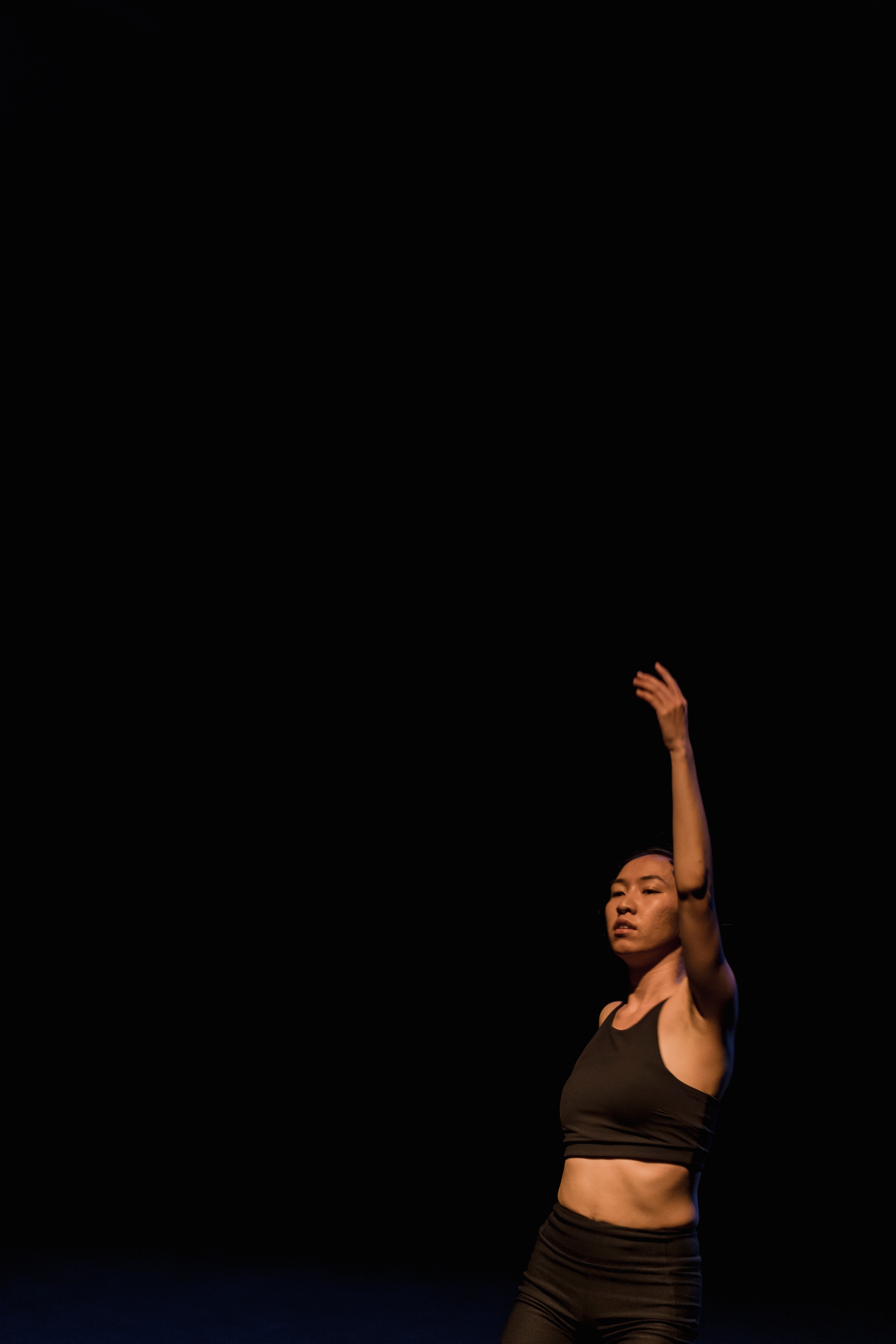 A portrait of dancer Lai Yi Ohlsen. She is seen against a dark background at the bottom right of the photo posing with left arm stretched directly above her head, as if mid-dance. 
