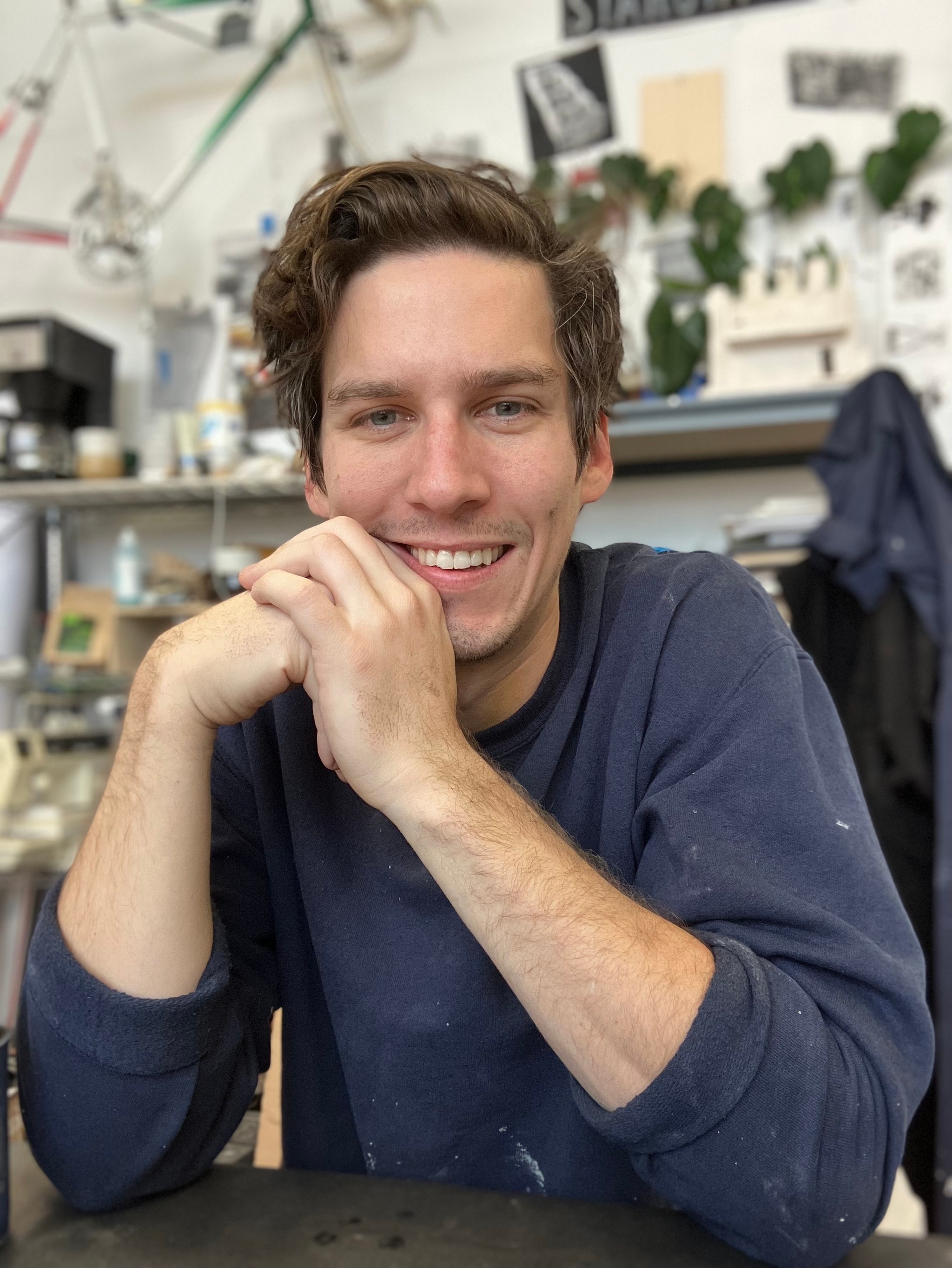 Artist Sean Starowitz, a white man with wavy brown hair, sits with his shoulders hunched and his hands brought together supporting his chin. He smiles and wears a blue sweatshirt. 