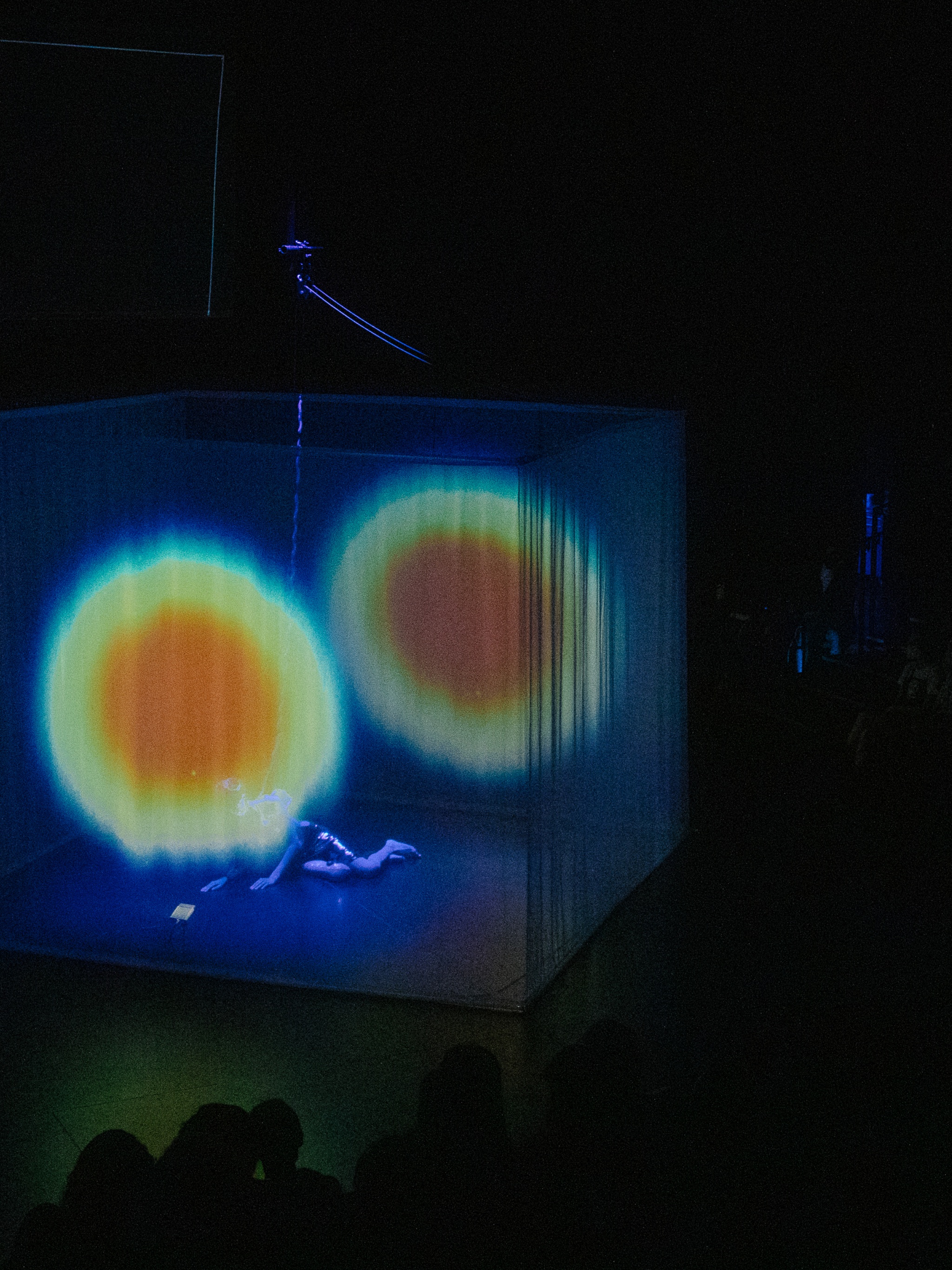 At the center of a darkened theater, sheer fabric hanging from the ceiling creates a cube on which a pulsating orange and green circle is projected. Through the fabric, a performer, the artist Yo-Yo Lin, can be seen wearing white lying on the floor.