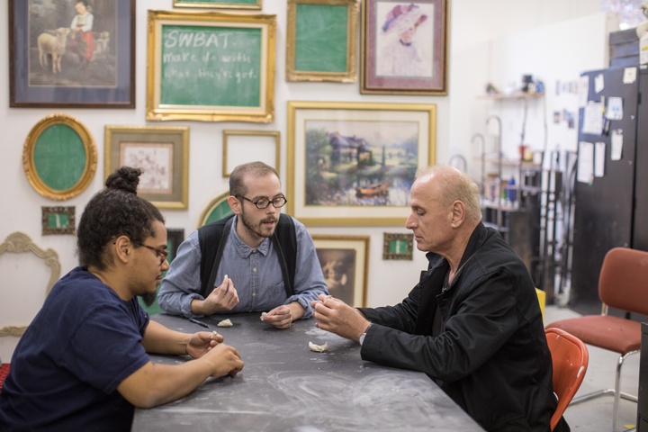 Three people sit at a table in a room hung with dozens of framed images. One person inspects a small white object while another leans toward them to explain something.