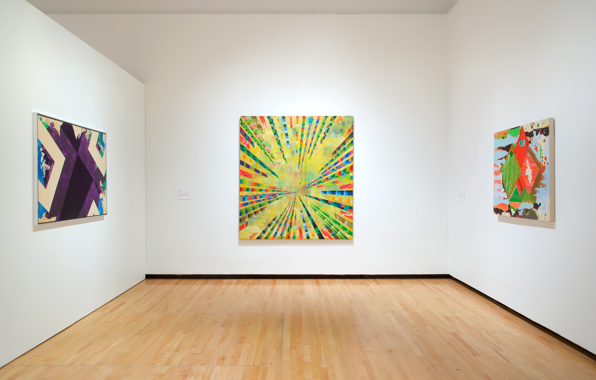 Three abstract, colorful paintings hang on white walls of a room with warm wood floors.