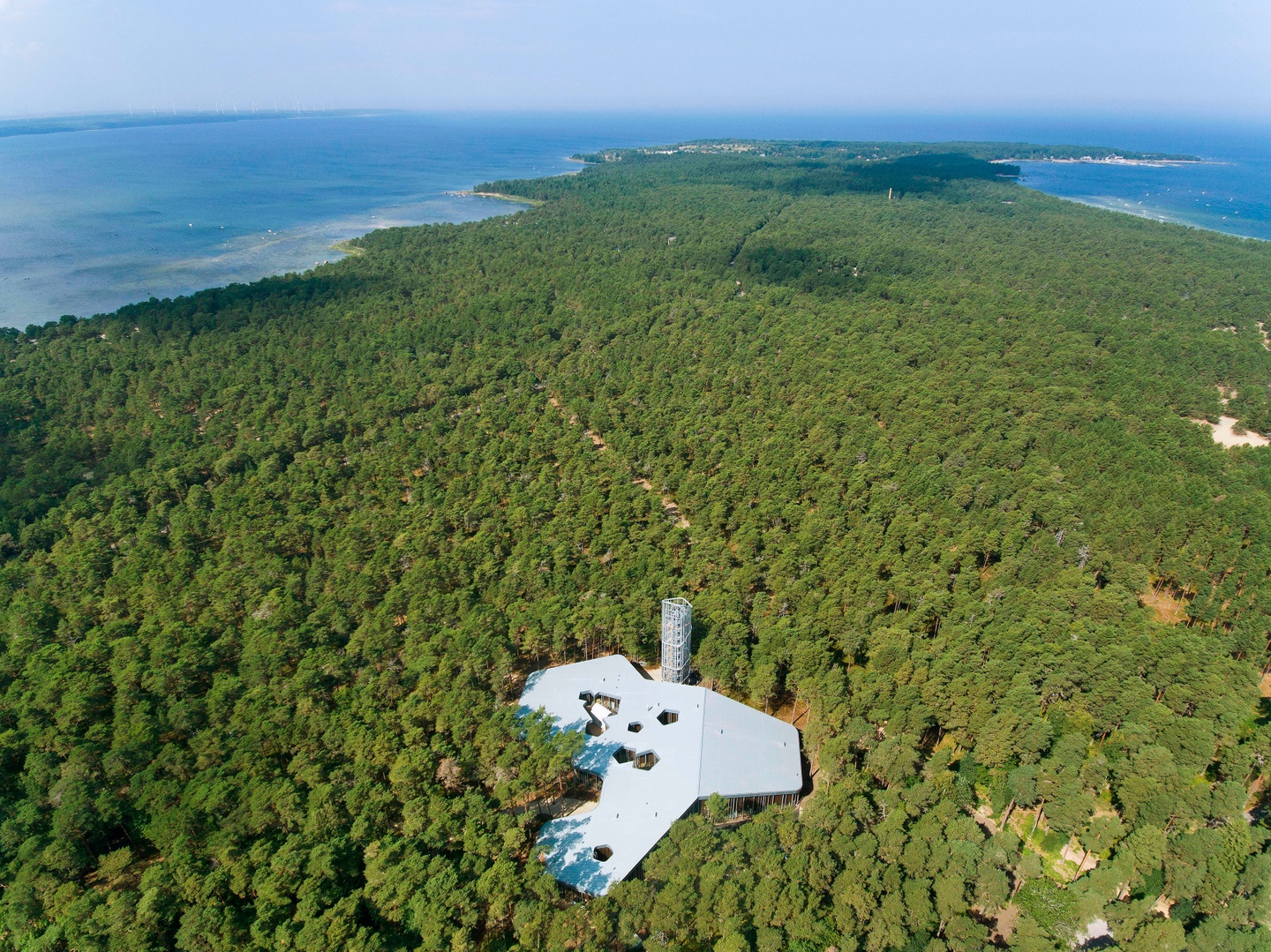 Aerial view of a dense, tree-filled area with water to the top of the photo. Near the bottom of the photo an irregularly shaped gray building with a flat roof and tower structure stands out.