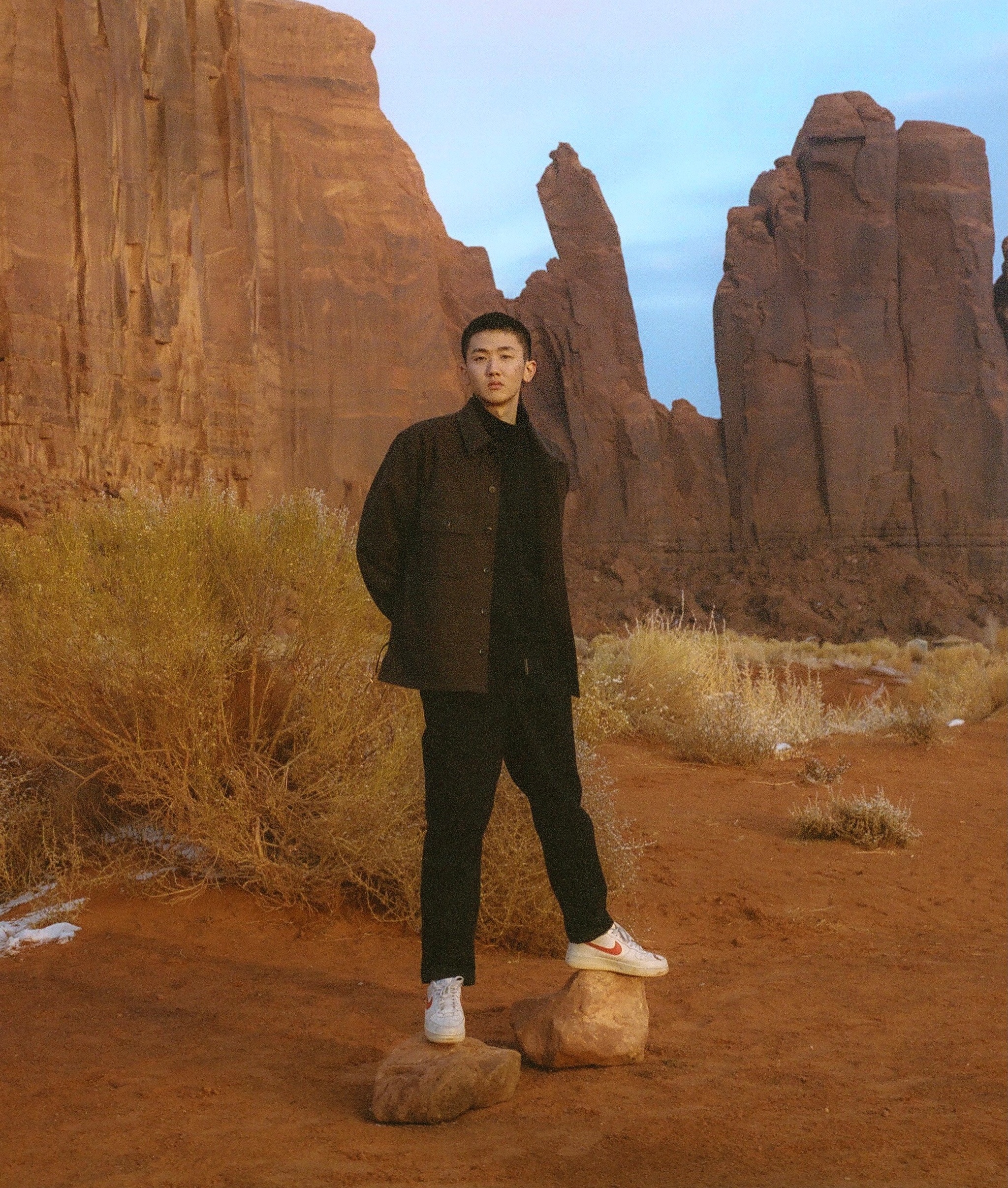 A young Chinese man perches on two rocks beneath his white sneakers, standing against a desert landscape with dusty cliffs rising up behind him. He wears black pants, a black shirt, and a dark green jacket. Between him and the cliffs are scrubby desert bushes. 