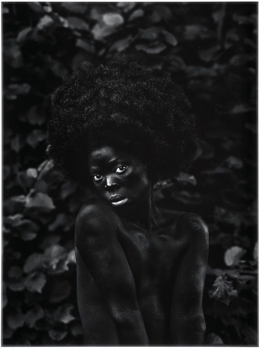 Black and white photograph of a person in the nude looking intensely at the camera and whose dark skin is emphasized.