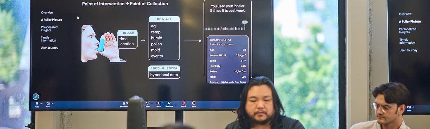 Two students sit in front of a tv monitor projecting their presentation to a group seated in front of them.