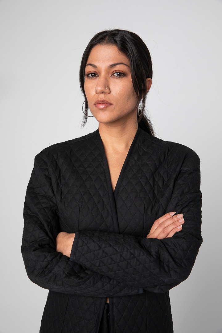 A woman in a black blazer crosses her hands over her chest. She stands against a blank white wall with her dark hair pulled back so only two strands fall on either side of her face.