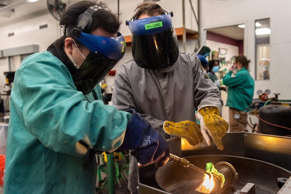 Two people in face shields and protective coats melt metal with a torch in a crucible.