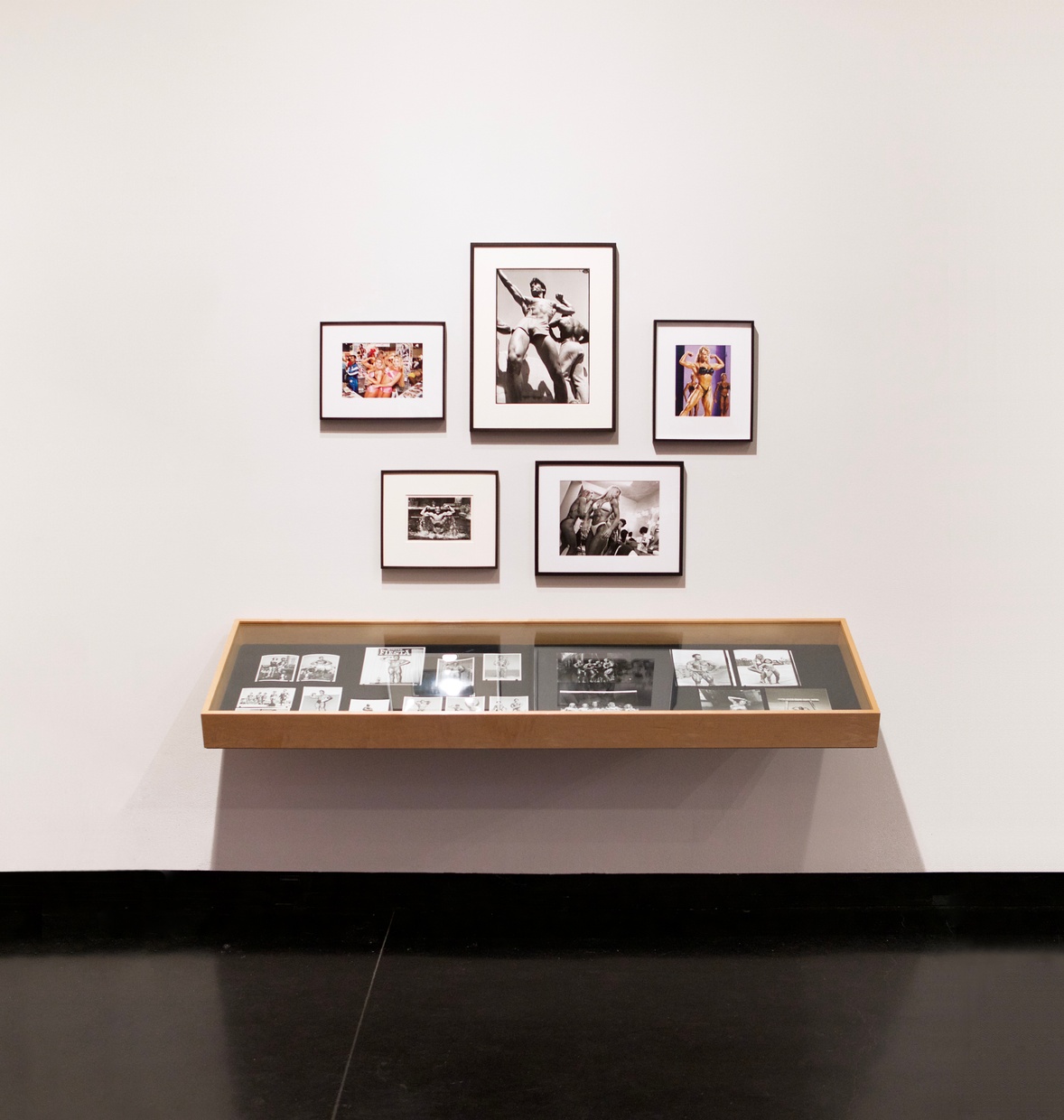 Five framed photographs featuring muscular bodies hung on a white wall above a glass case with black and white photographs.