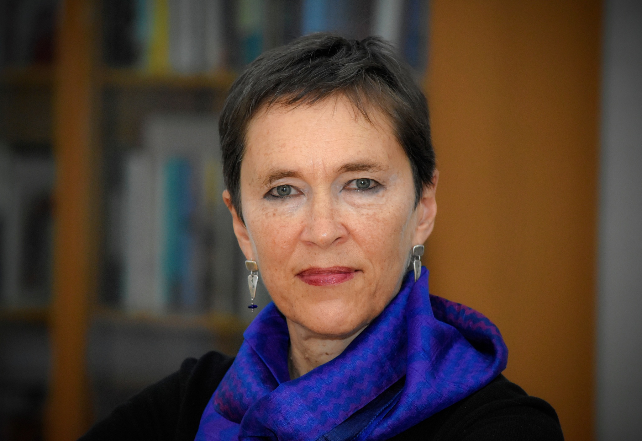 A white woman with short cropped brown hair with a shelf of books in the background. She wears a royal blue scarf around her neck. 