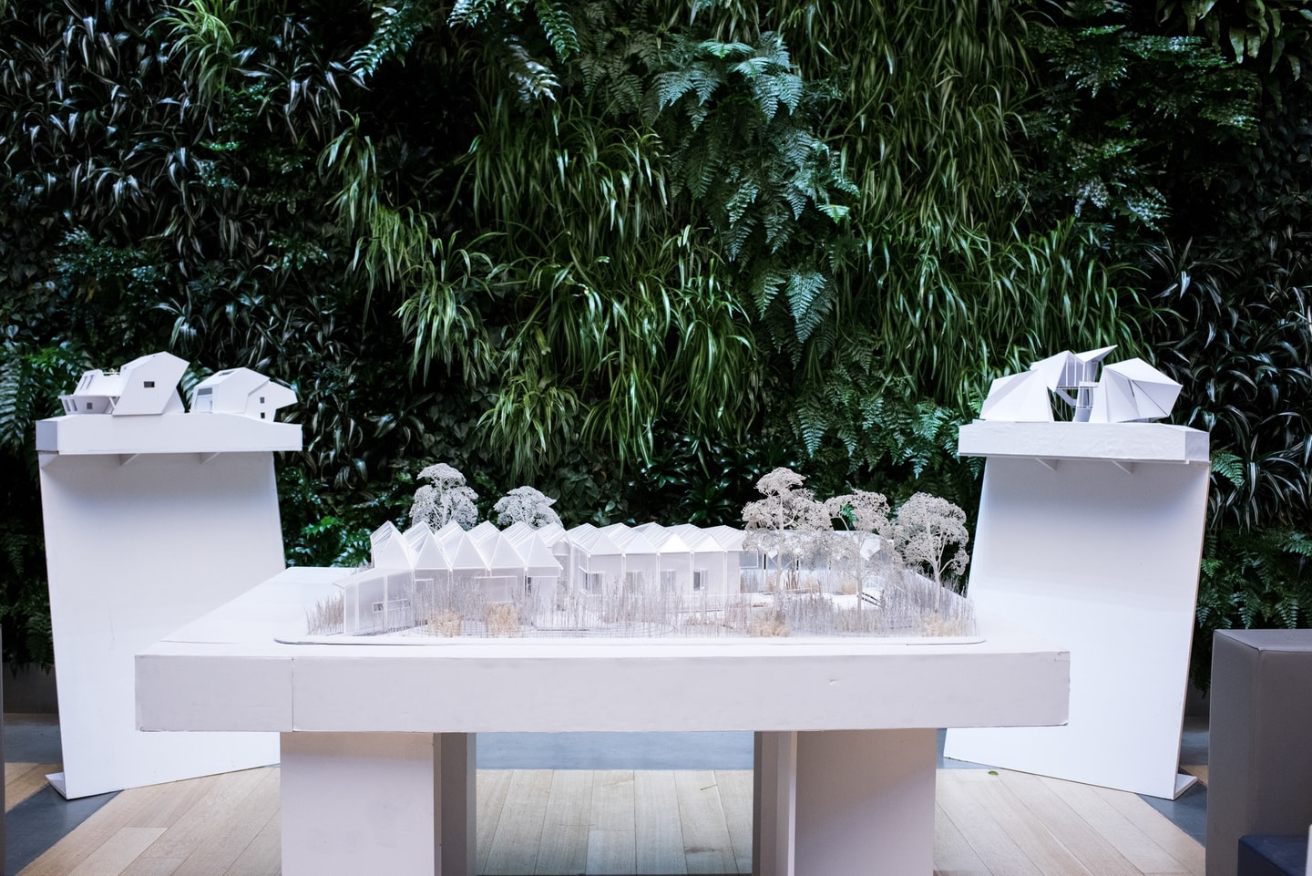 Three white architectural models on stands in front of a living green wall.