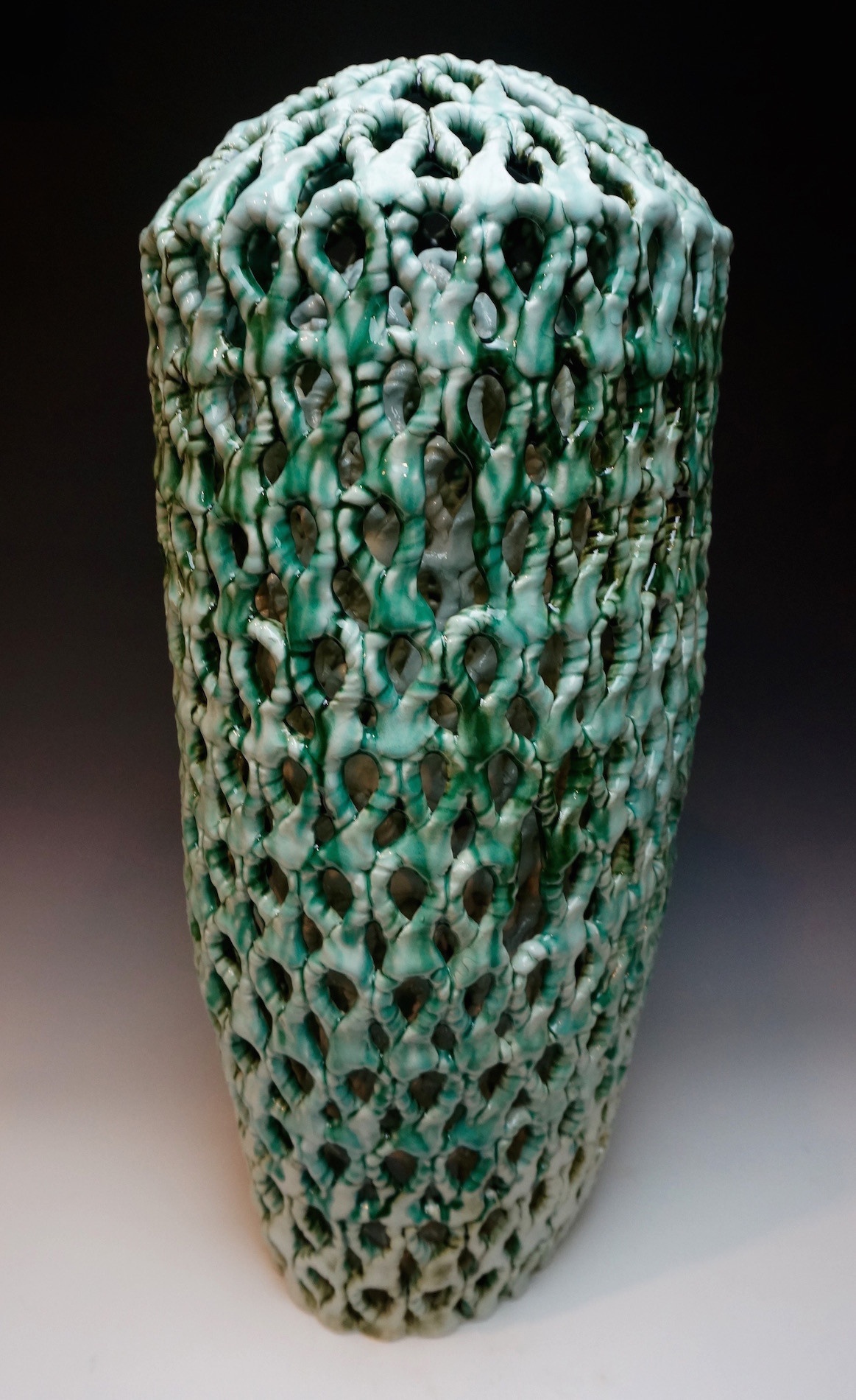 An intricately looped/patterned vessel, the top/mouth of it closed off — its surfaces are a vibrant emerald hue streaked with white.