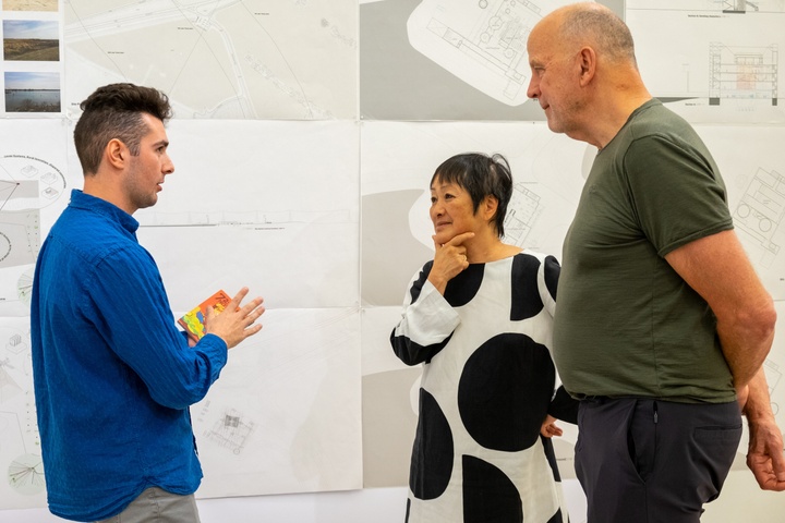 A student converses with architects Billie Tsien and Tod Williams, standing in front of his work pinned up on the wall.