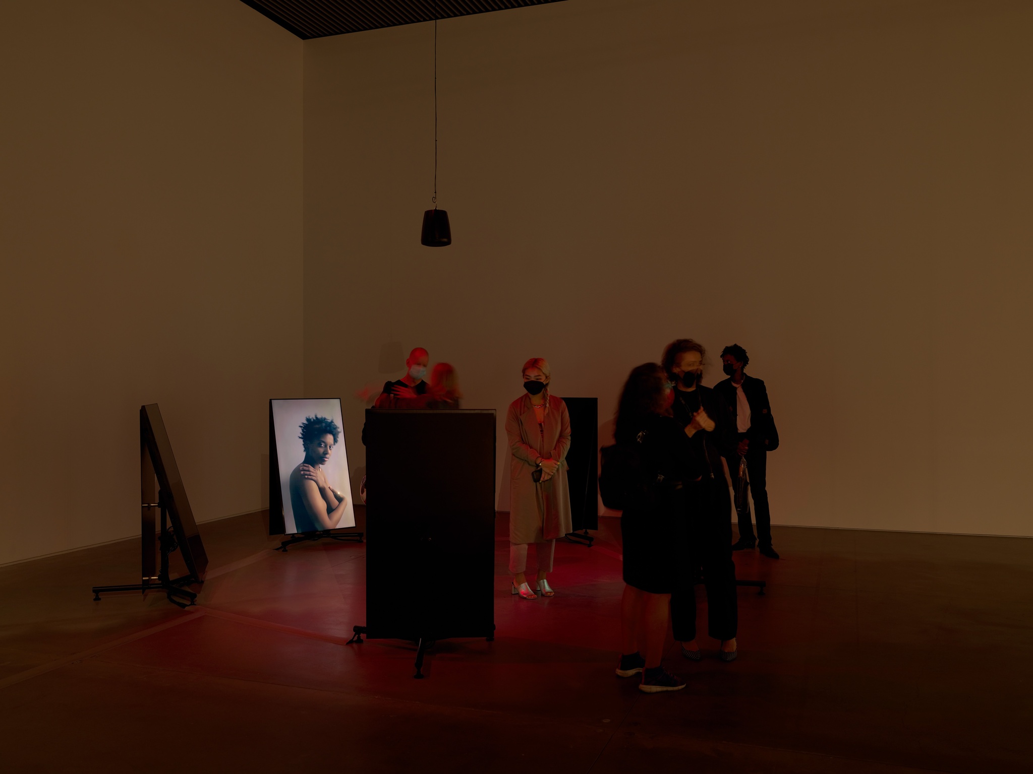 An art gallery with dim lighting and a group of people standing outside and within a circle of five vertical standing video screens. One of the screens shows a Black woman in profile looking out at viewers.