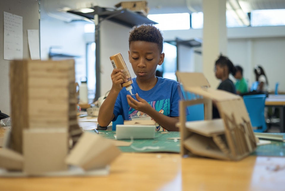 A middle school student in a blue shirt is sitting at a table using glue on his architecture project.