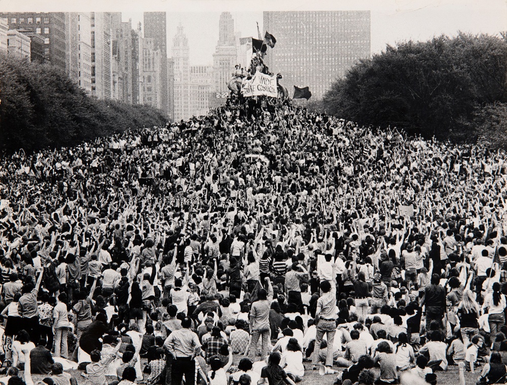 A black and white photograph shows a city park filled with thousands of people facing a large statue in the center draped with flags and banners. The figures have their fists raised in the air.