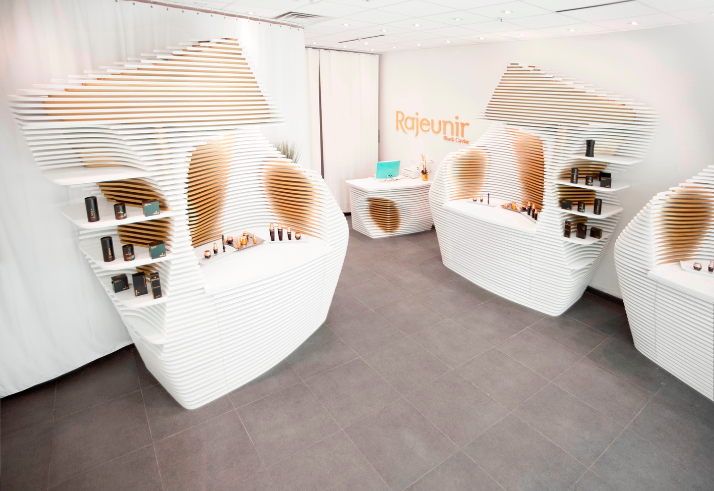 Interior of a white space with a concrete floor and three white display shelves formed of layered boards with divots cut out of them to hold various bottles and small containers. Gold letters on the wall read "Rajeunir."